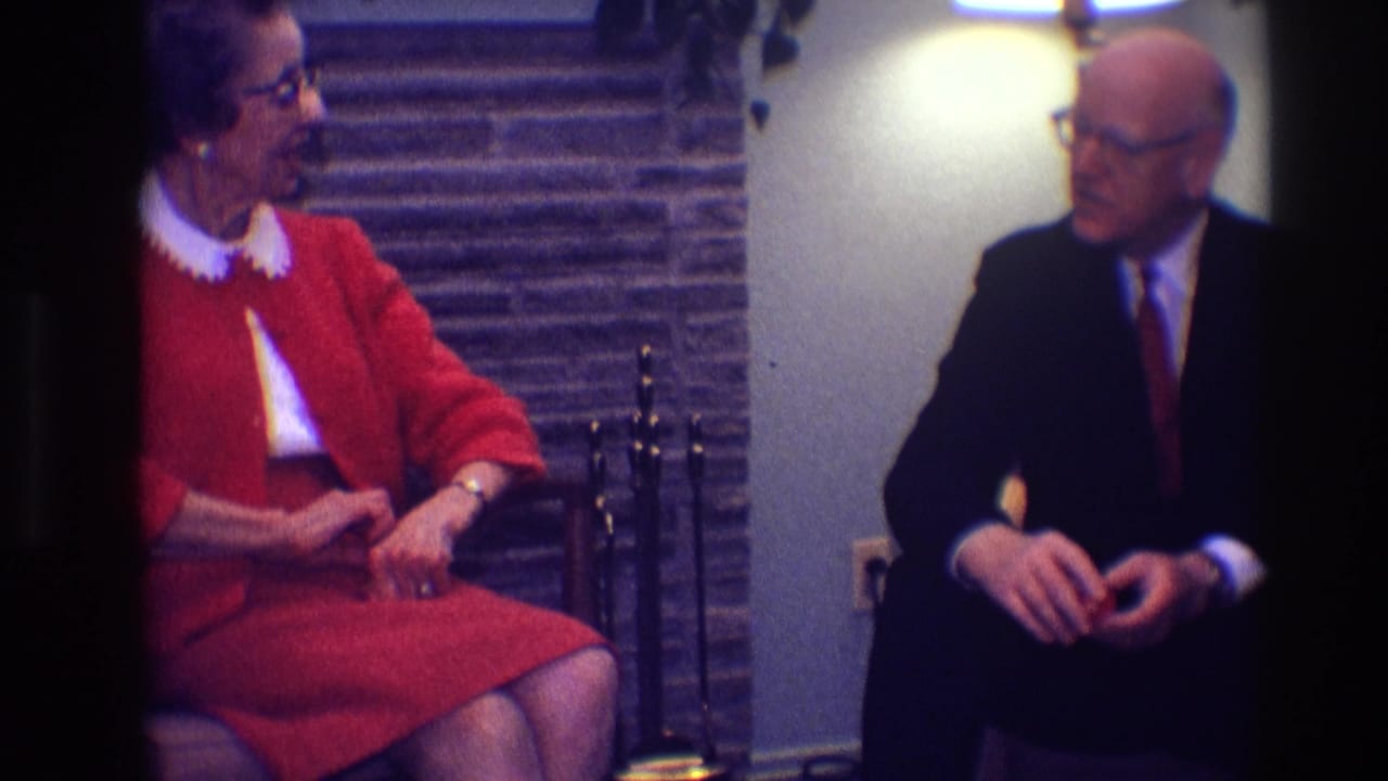 Elderly woman in red dress sits beside a suited man in a living room.
