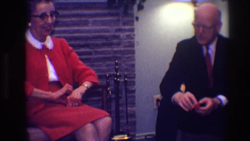 1968 Toledo frame with an elderly woman in red and a man in a dark suit seated in a private living room.