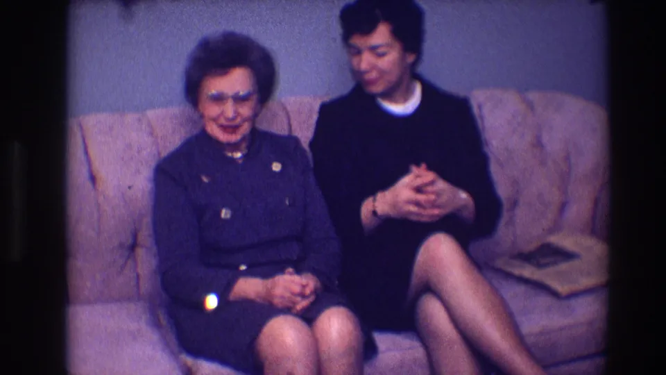 1968 Toledo frame of an older woman in dark clothing and a younger woman seated side by side on a couch.
