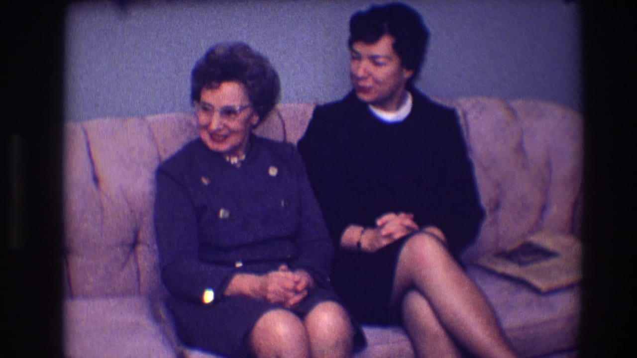 Two adult women sit side by side on a sofa while looking down and toward each other.