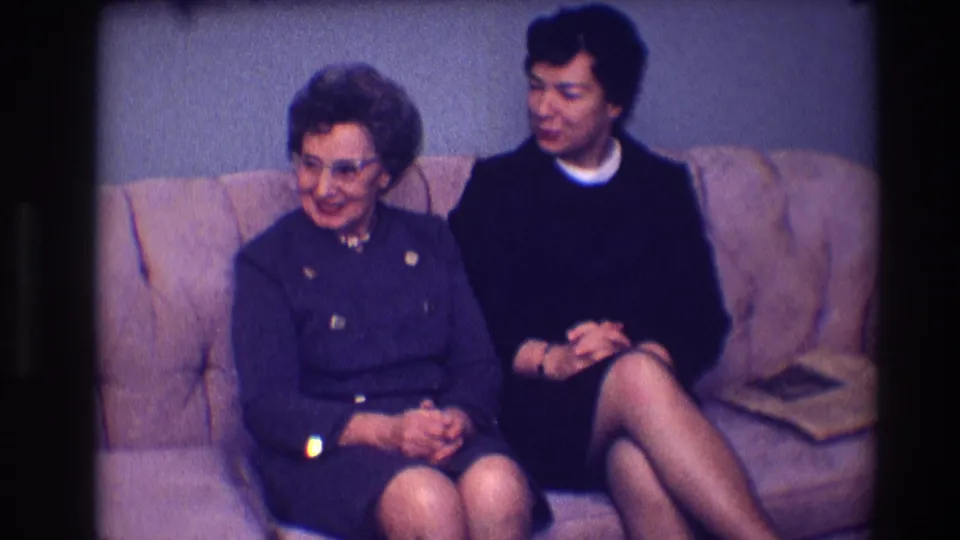 The older woman in glasses and the younger woman in a dark dress sit on a brown sofa during a quiet 1968 Toledo domestic scene.