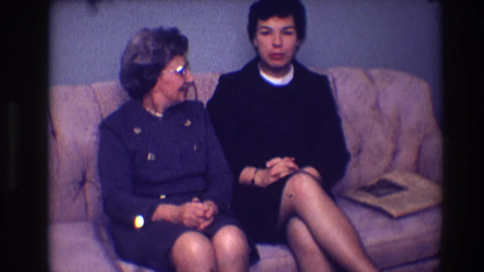 In a 1968 Toledo living-room scene, the older woman in glasses turns toward the younger woman seated beside her on the sofa.