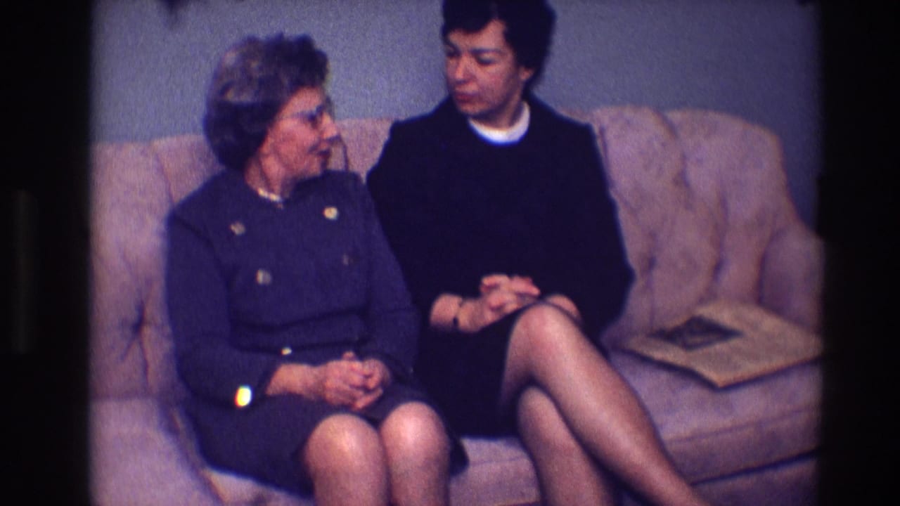 Two women sit on a tan sofa in a blue-walled room, looking toward each other.