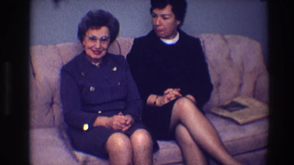 The older woman in glasses faces the camera while the younger woman sits beside her on a tan sofa in the 1968 interior.