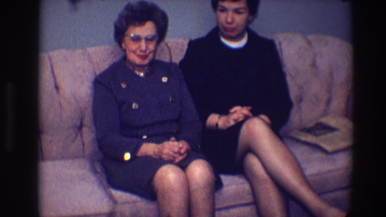Two women sit on a couch as the older woman faces forward and the younger woman looks down.