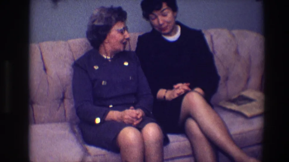 1968 Toledo home-movie frame of two women facing each other in conversation on a light-colored couch.