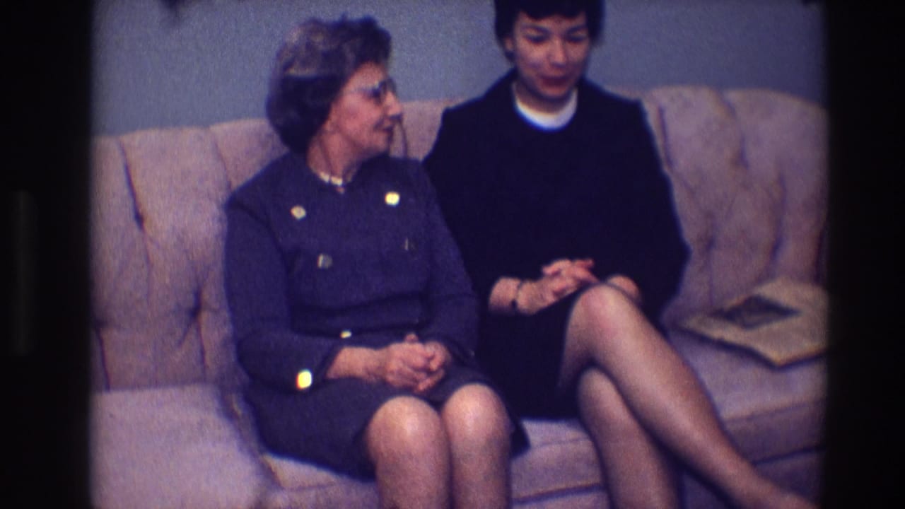 Two women sit on a tan sofa, one in a blue suit and one in a black dress.