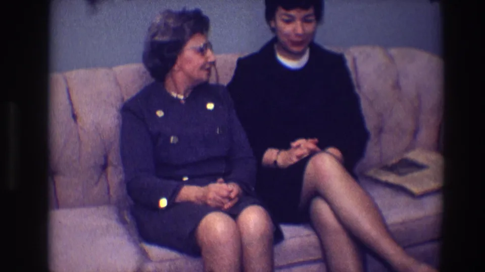 In a Toledo, Ohio home-movie frame from 1968, two adult women sit on a tan sofa during an indoor conversation.