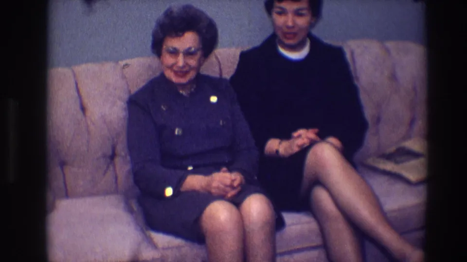The 1968 Toledo home-movie frame shows an older woman in glasses smiling beside a younger woman on a tan sofa.