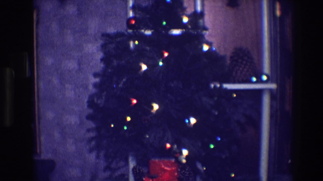 Dim indoor Christmas tree with colored lights, pinecones, and a red gift box.