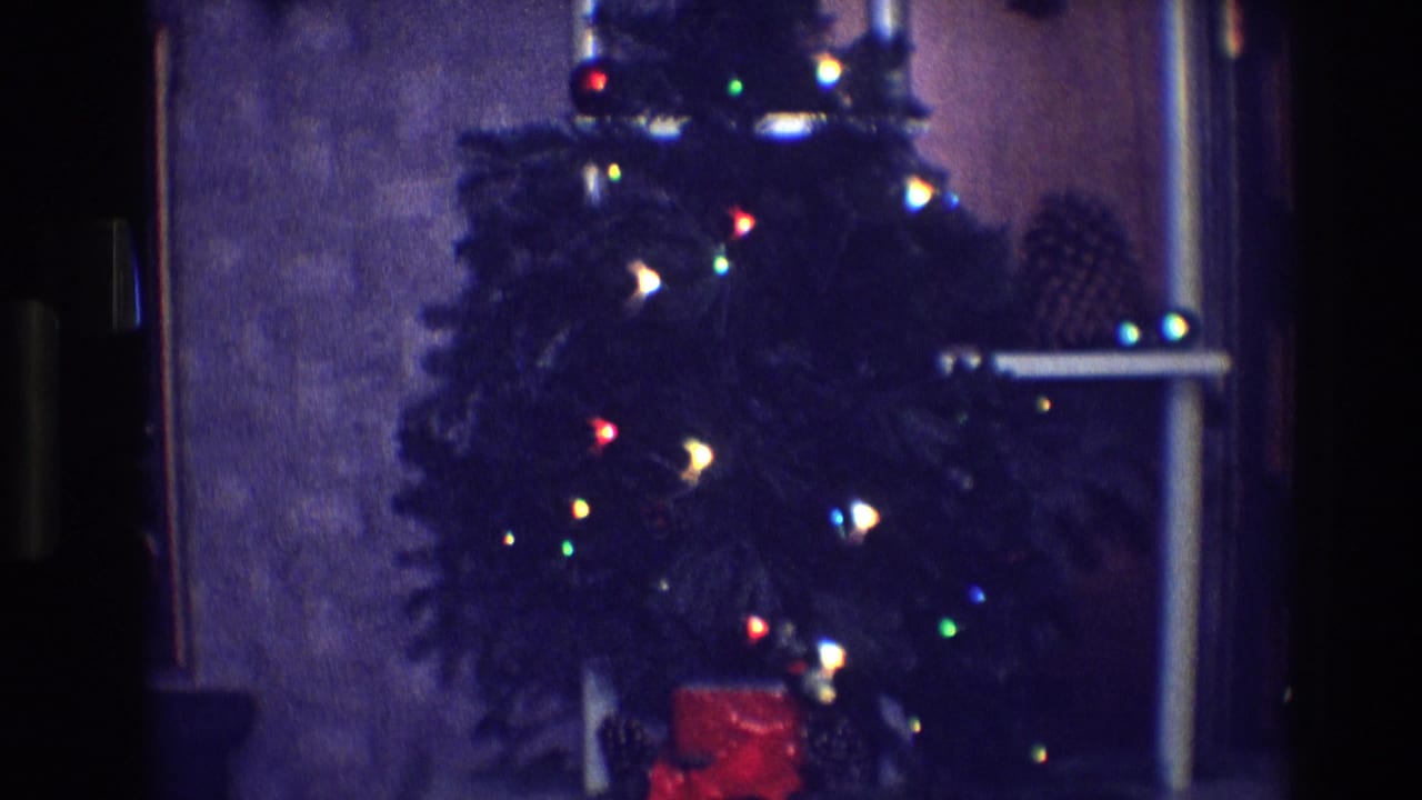 Lit Christmas tree with colored bulbs, pinecones, and a red gift near a window.
