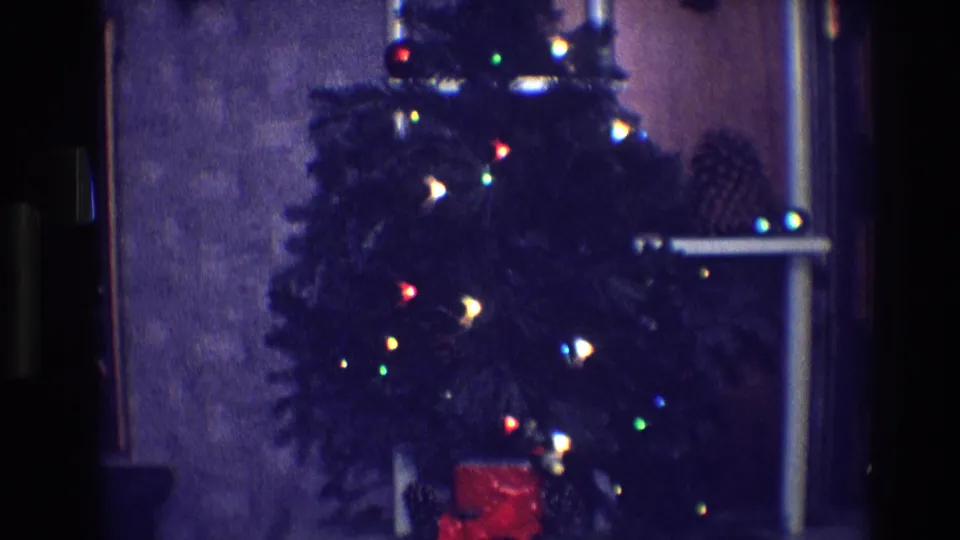 1968 Toledo home-movie frame showing a small indoor Christmas tree with multicolored lights, pinecones, and a red gift below.