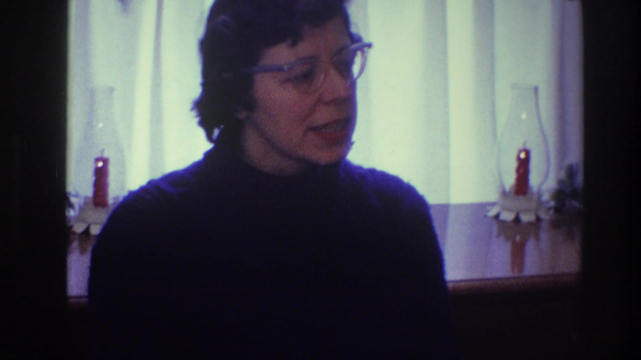 Adult woman in glasses and a dark turtleneck speaking near a curtained window.