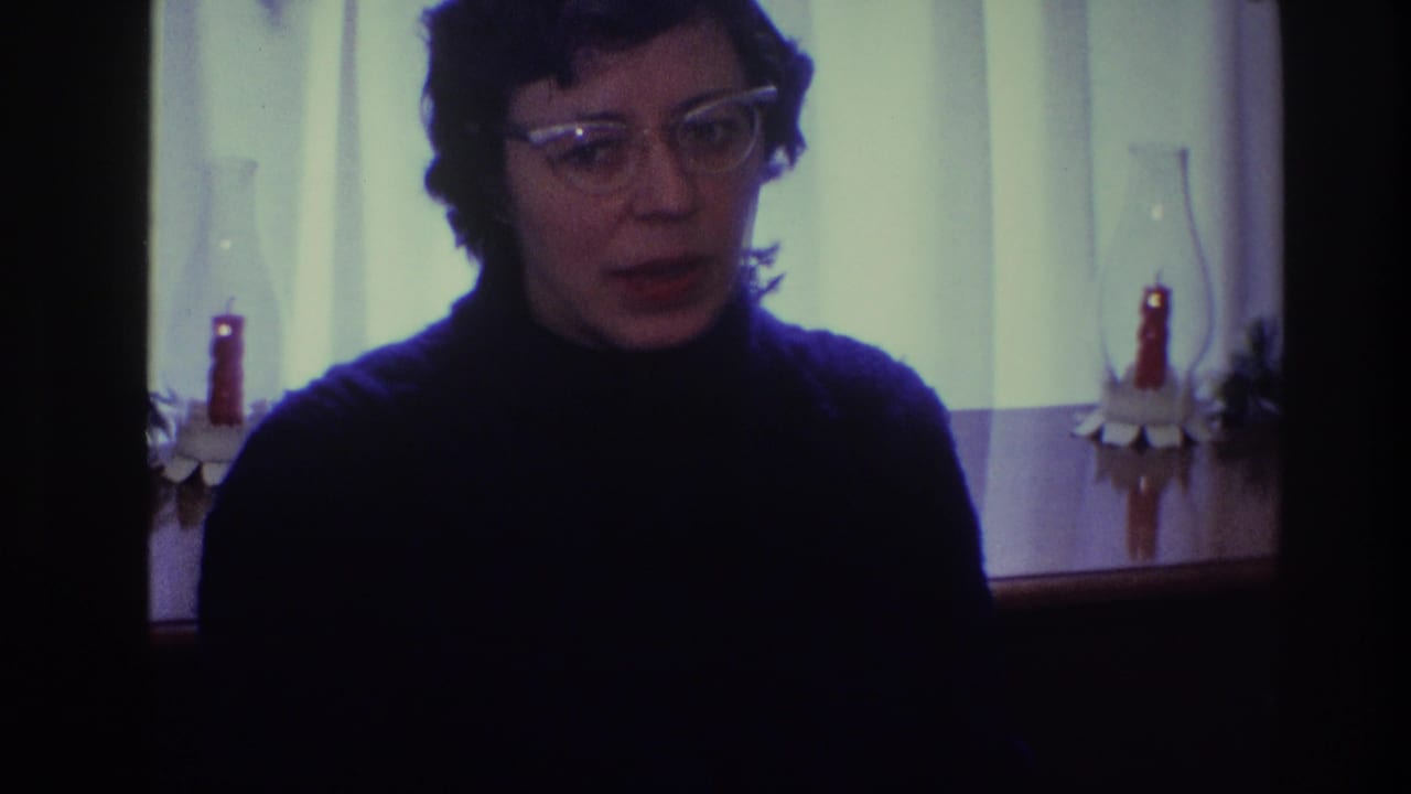Adult woman in glasses faces forward in a dim interior with candles behind her.