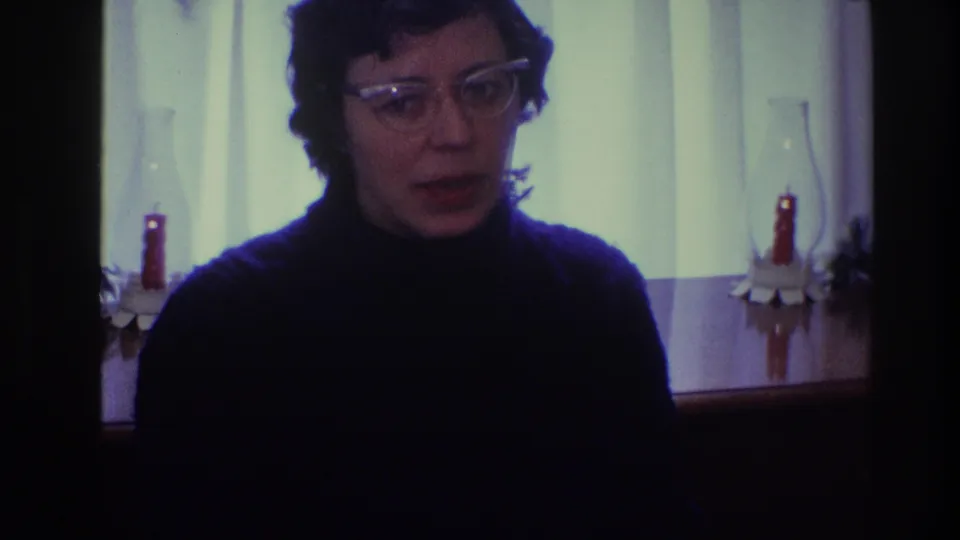 The woman faces toward the camera in the Toledo 1968 home-movie interior, with curtained light and red candles visible behind her.