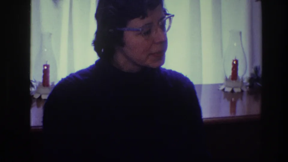The woman turns in profile or three-quarter view in this Toledo 1968 home-movie frame, with red candles and soft window light behind her.