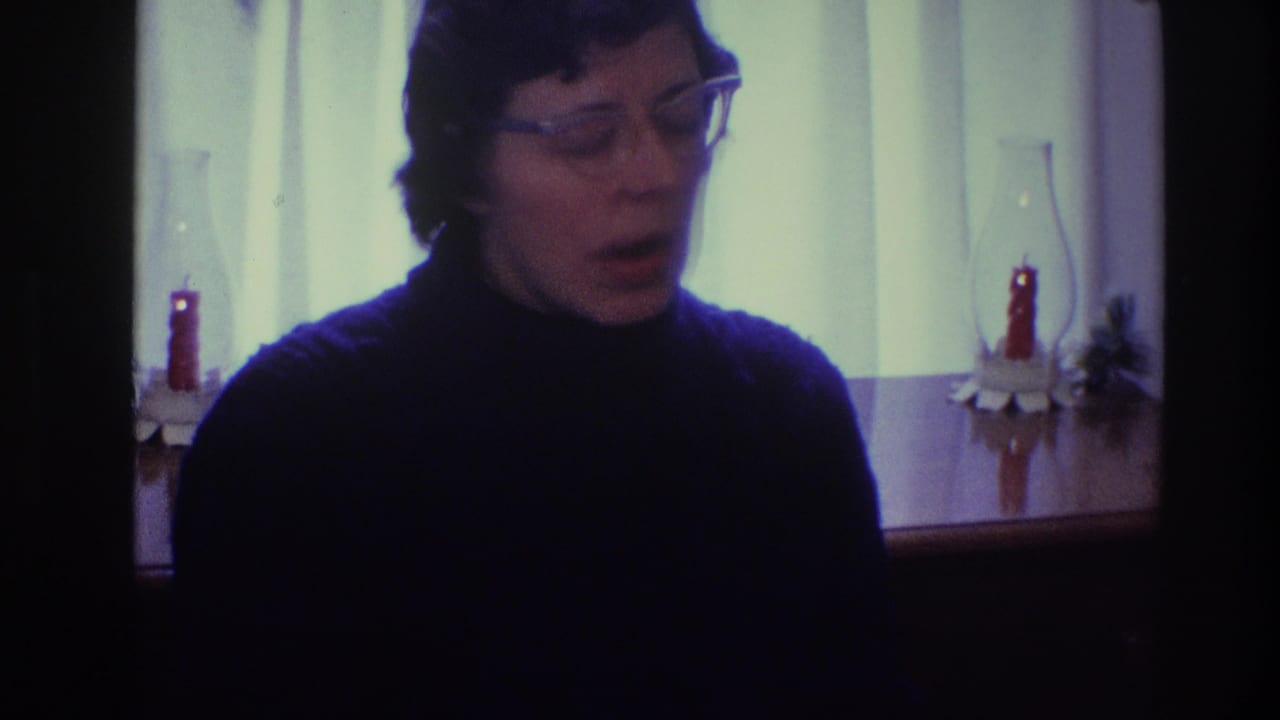 Woman in glasses and a dark turtleneck speaks in front of a curtained window.