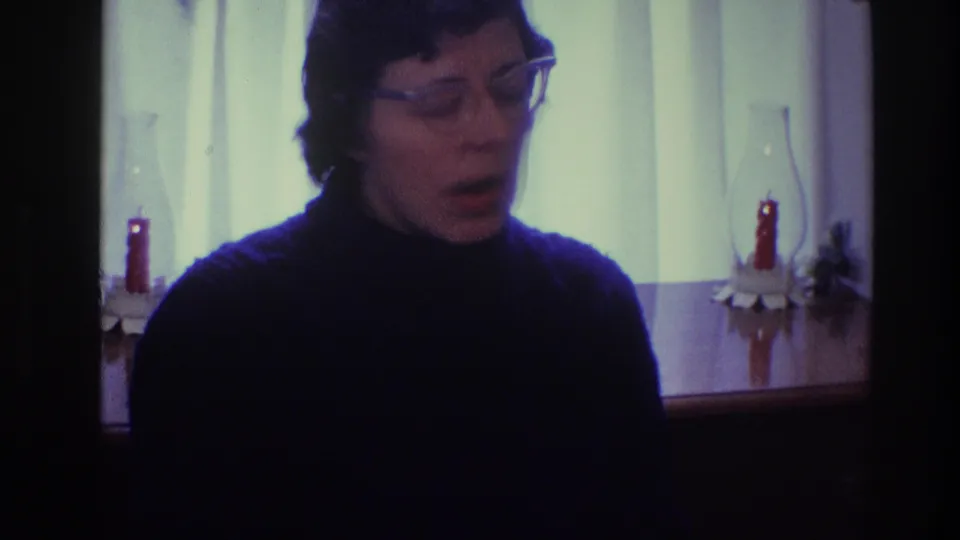 In a 1968 Toledo home interior, a woman with clear-framed glasses faces the camera near candles and a curtained window.