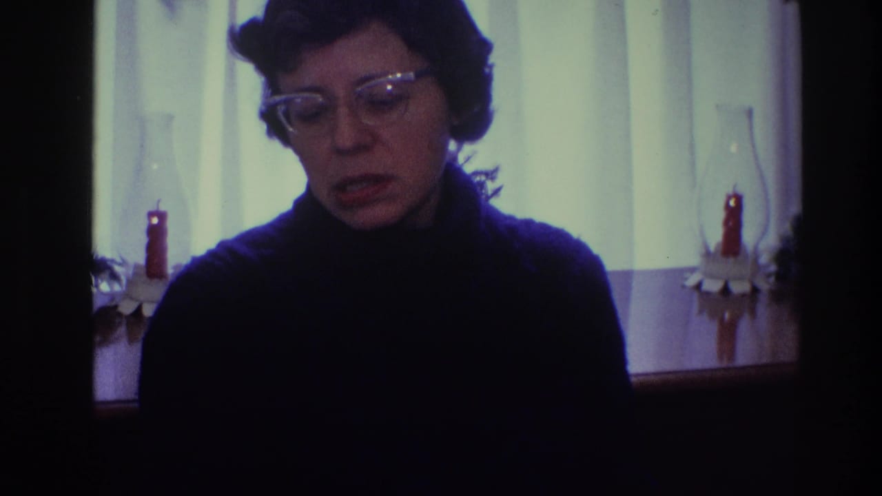 Woman with glasses looks downward while seated in a dim home setting.