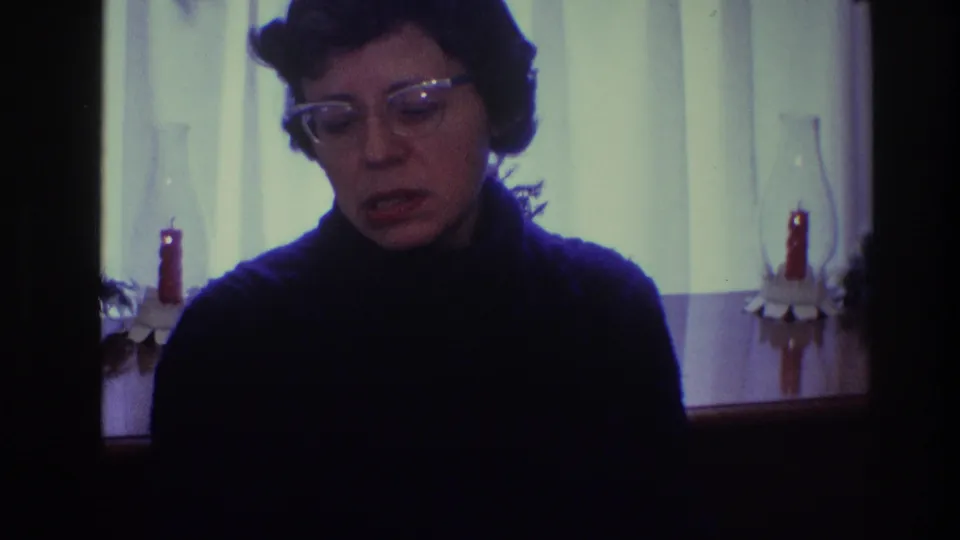 A woman in a dark turtleneck appears to speak or pause in a 1968 Toledo home, with candles and window curtains behind her.