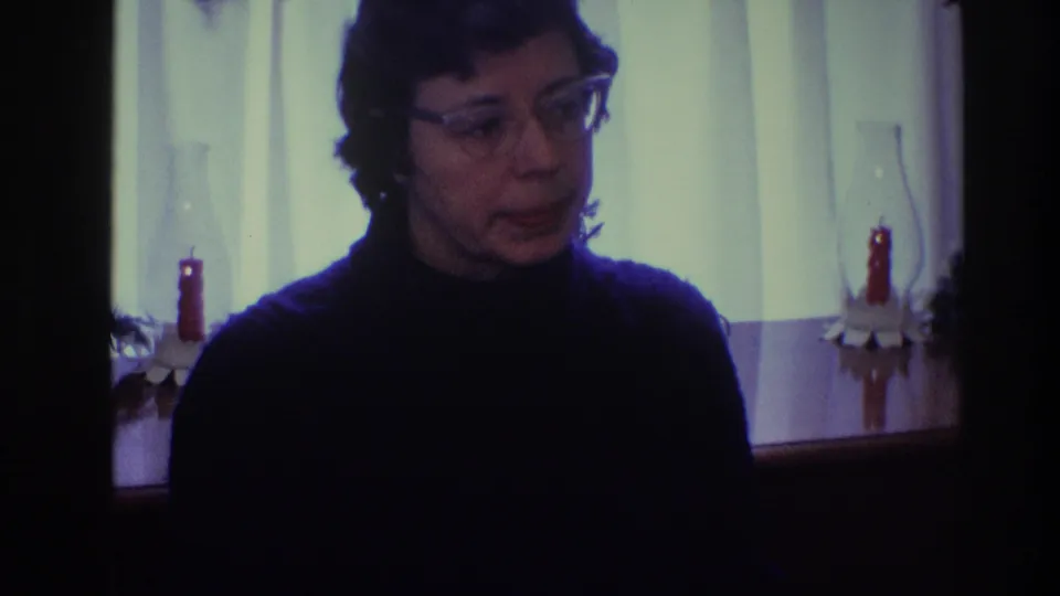 A woman in clear-framed glasses turns slightly in a 1968 Toledo home-movie frame, with red candles visible behind her.
