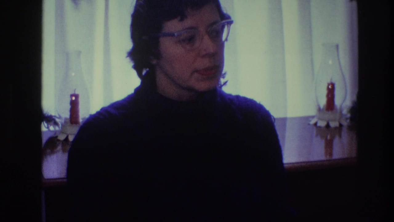 Woman in glasses beside a curtained window and candle holders in a dim home interior.