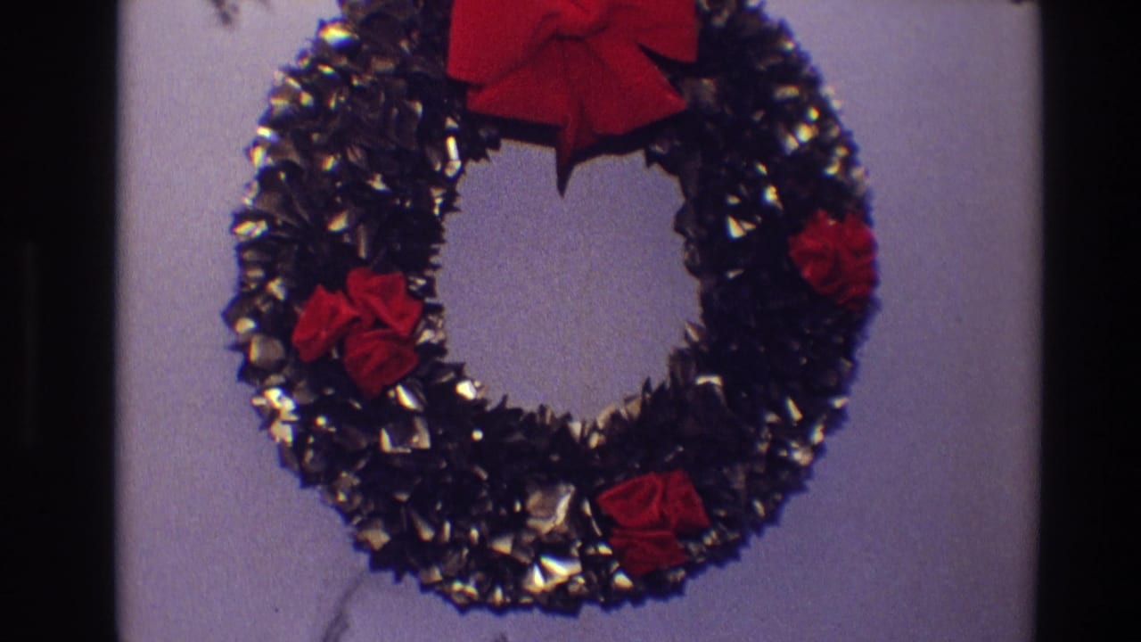 Close view of a dark tinsel Christmas wreath with red bow and fabric flowers on a pale wall.