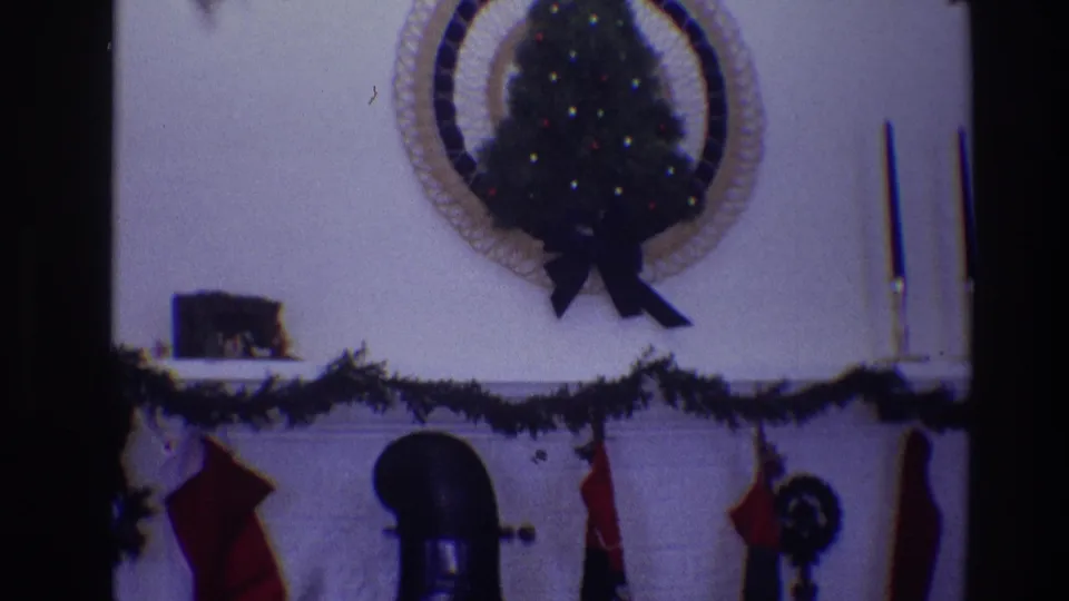 1968 Toledo home-movie frame showing a Christmas mantel with garland, red stockings, taper candles, and a small lit tree above a white fireplace.