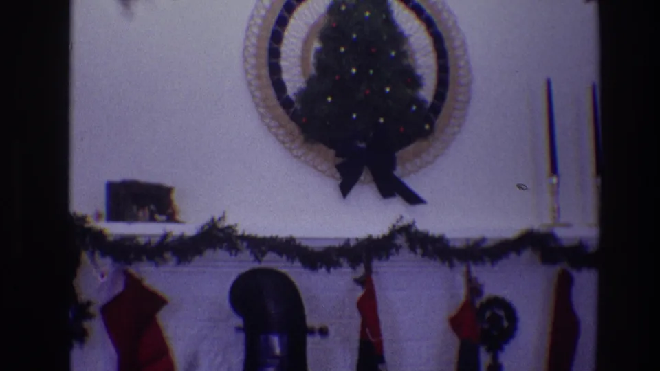 1968 Toledo 8mm frame of a decorated Christmas mantel with a small tree in a circular frame, red stockings, garland, and candles.