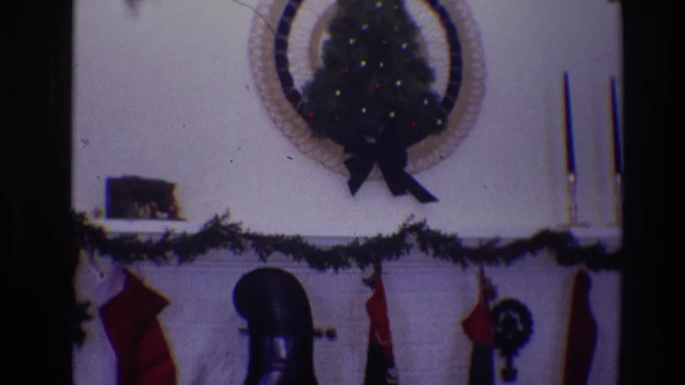 1968 Toledo 8mm frame of a private home Christmas mantel, including garland, stockings, a wall-mounted tree display, and candles.