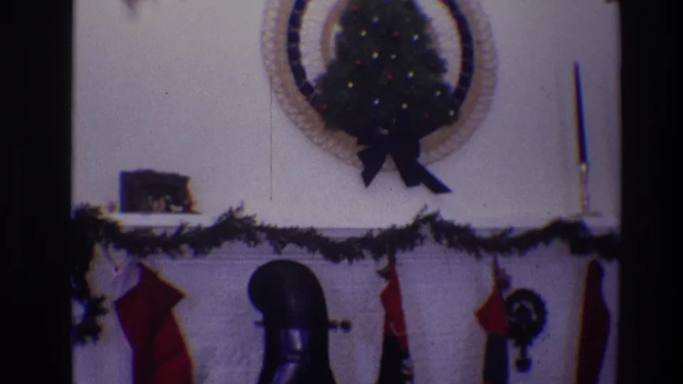 1968 Toledo frame showing a white fireplace mantel dressed with garland and stockings below a small decorated wall tree.
