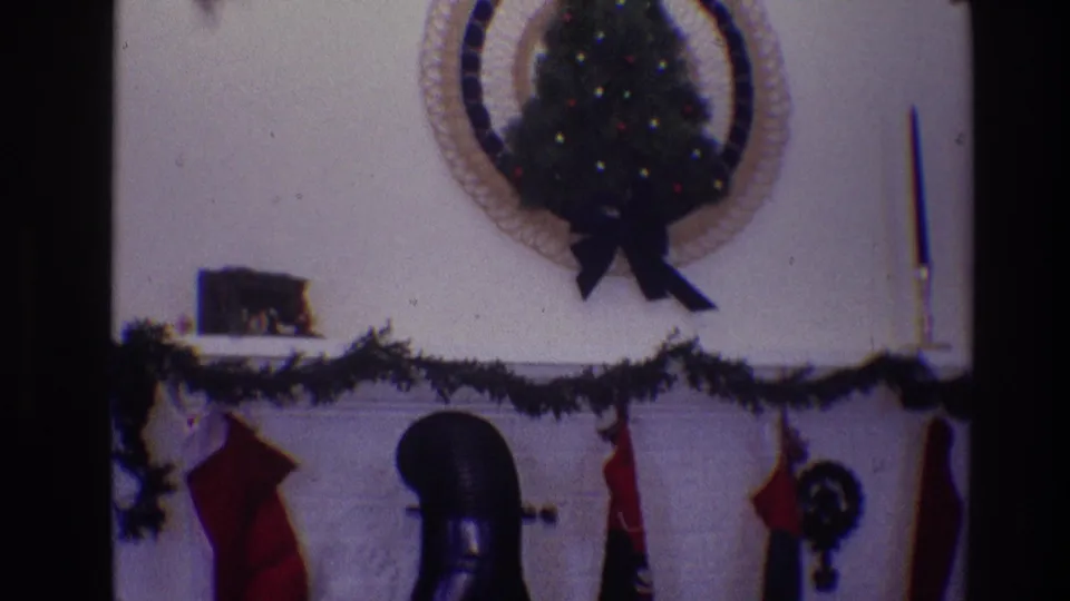 Soft 1968 Toledo home-movie view of a Christmas fireplace mantel with dark garland, hanging stockings, and a small decorated wall tree.