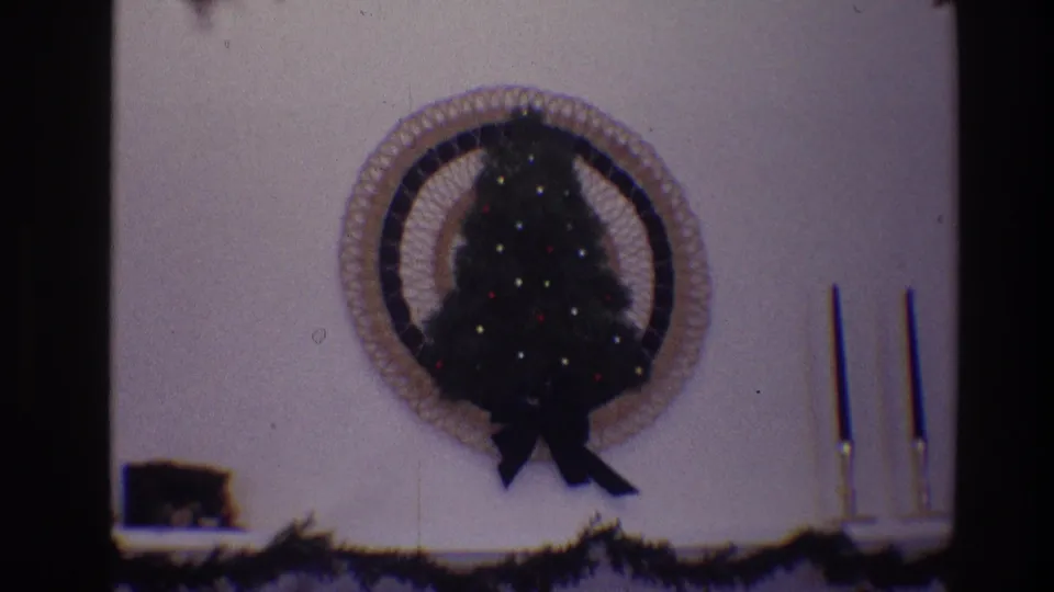 1968 Toledo home-movie frame showing a small lighted Christmas tree in a circular woven wall frame, with garland and candlesticks nearby.