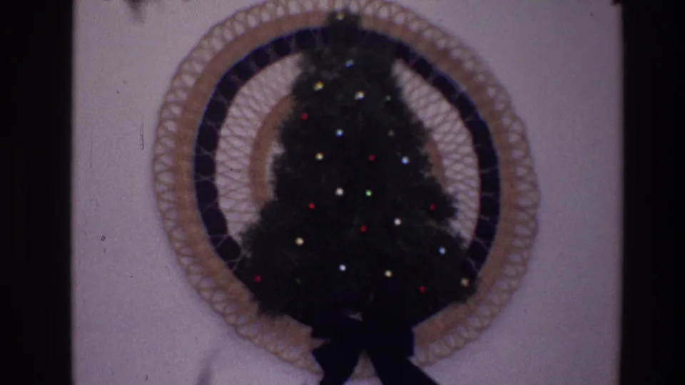 Close 1968 Toledo frame of a small artificial Christmas tree with colored lights set within a circular woven wall decoration.