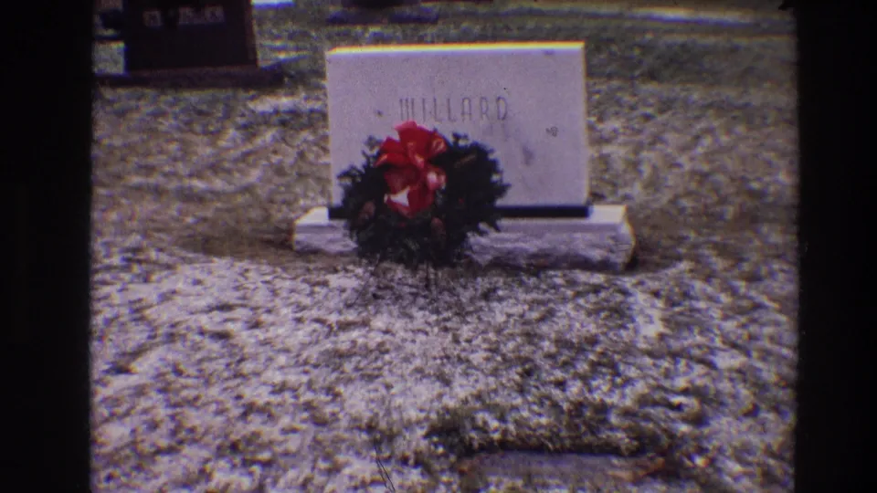 1968 Toledo home-movie frame showing a Millard grave marker with a red-bowed wreath on gravel cemetery ground.