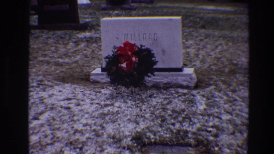 1968 Toledo home-movie frame with the wreath partly obscuring the MILLARD grave marker on icy cemetery grass.