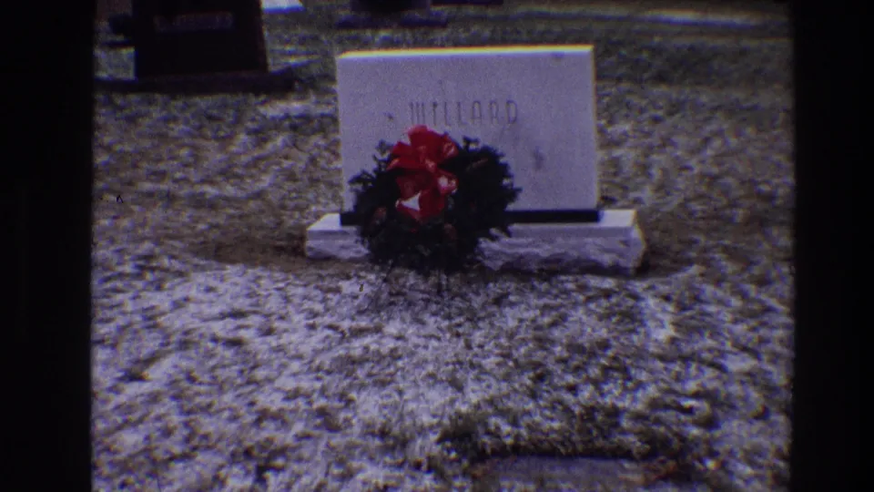 Final frame holds on the MILLARD headstone, wreath, and bright red bow, with neighboring cemetery markers faintly visible behind.