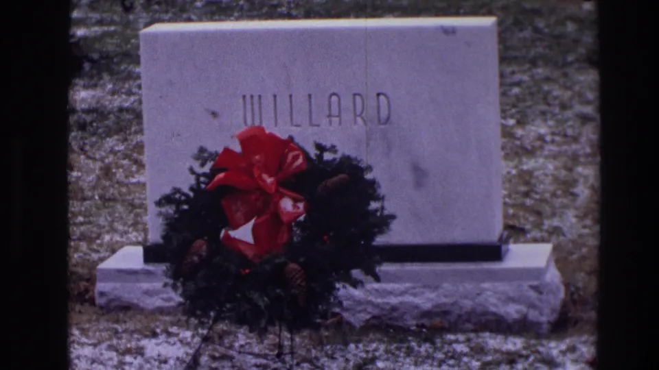 1968 Toledo home-movie frame of a gray WILLARD tombstone, red-bowed wreath, stone base, and light snow in a cemetery.