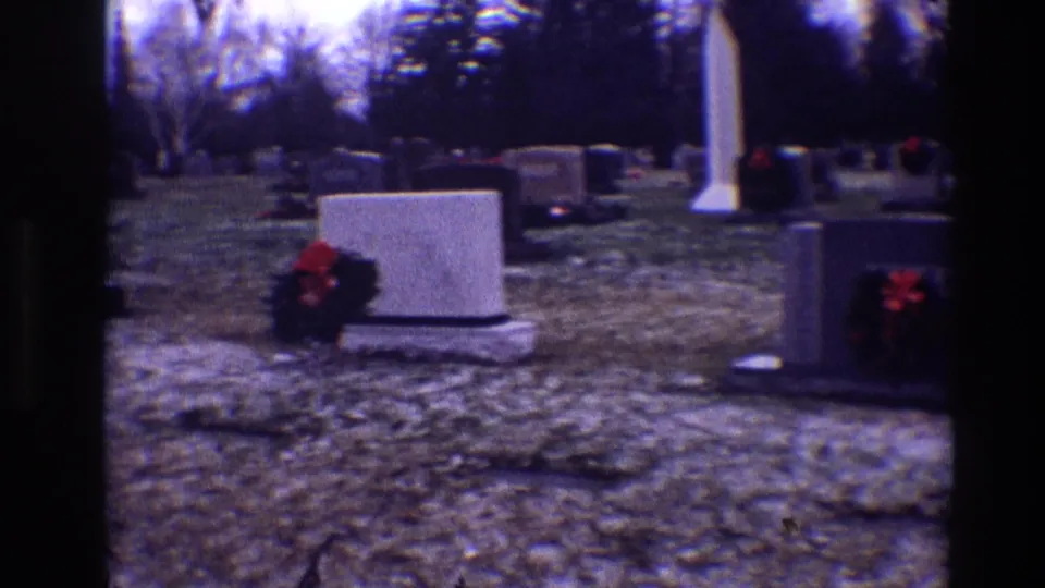1968 Toledo cemetery frame showing wreaths on gravestones, a tall white monument, bare trees, and snow-dusted grass.