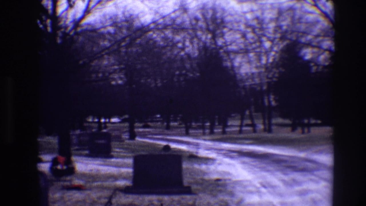 Snow-dusted cemetery path with grey headstones and bare winter trees.