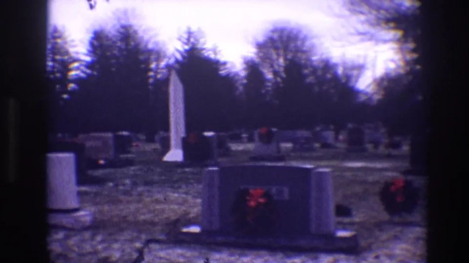 1968 Toledo cemetery view with a tall white monument, low grey tombstones, red wreaths, bare trees, and snow-dusted ground.