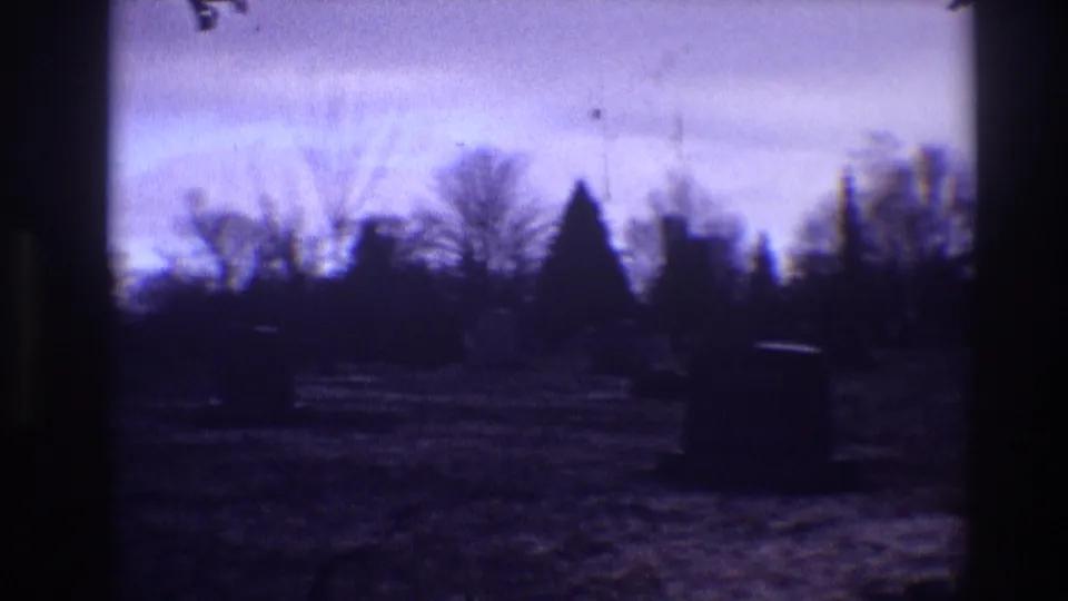 1968 Toledo home-movie frame showing silhouetted grave markers and bare trees under a blue-purple dusk sky.