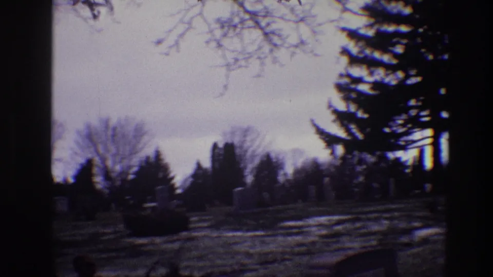 1968 Toledo, Ohio 8mm frame showing a winter cemetery with bare trees, evergreens, scattered headstones, and a dark foreground trunk.