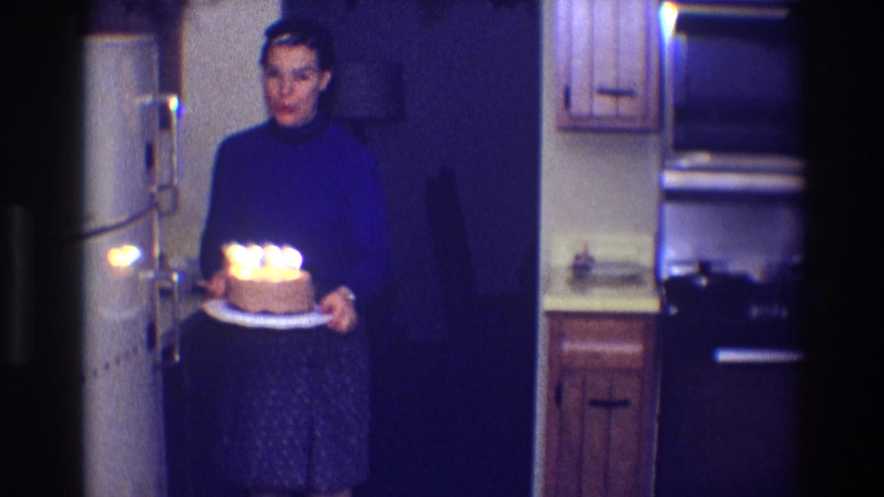 Woman carries a small birthday cake with lit candles through a dim 1968 Toledo kitchen.