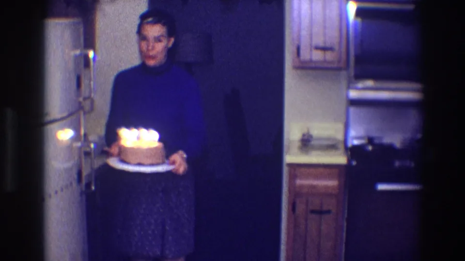 Woman carries a small birthday cake with lit candles through a dim 1968 Toledo kitchen.
