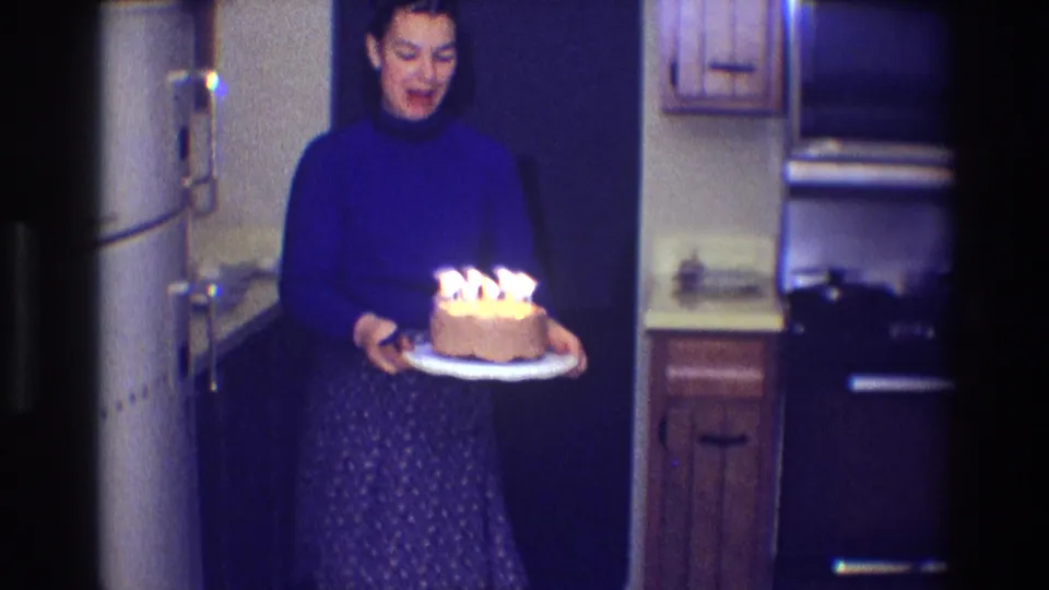 Woman holds a candlelit birthday cake in a private kitchen, with appliances and cabinets behind her.