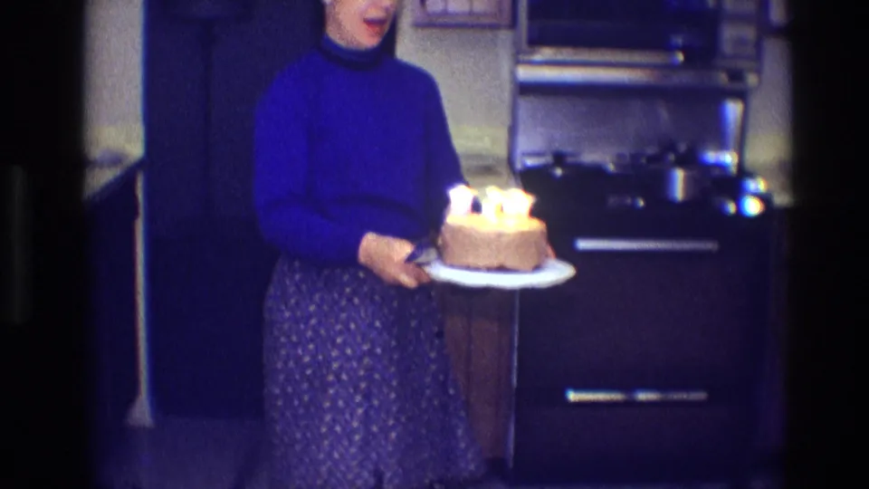 Closer view of the woman carrying the candlelit cake past the stove in the Toledo kitchen.