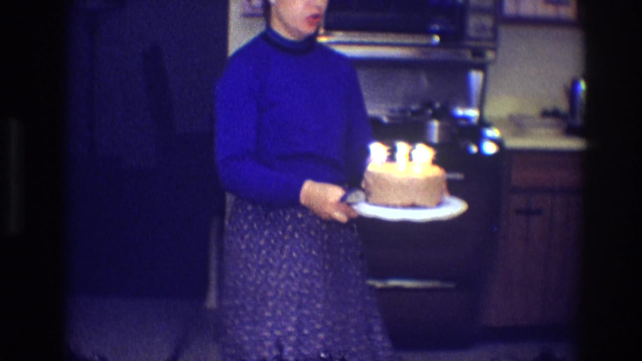 Woman in blue turtleneck holds a birthday cake with lit candles in a kitchen.