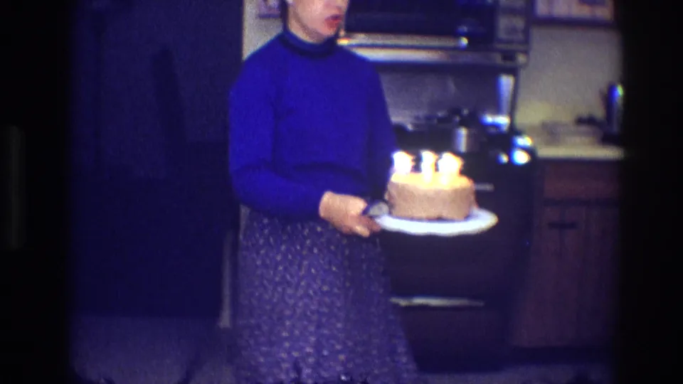 In a 1968 Toledo home kitchen, a woman in a blue turtleneck and patterned skirt holds a lit birthday cake near wood cabinets.