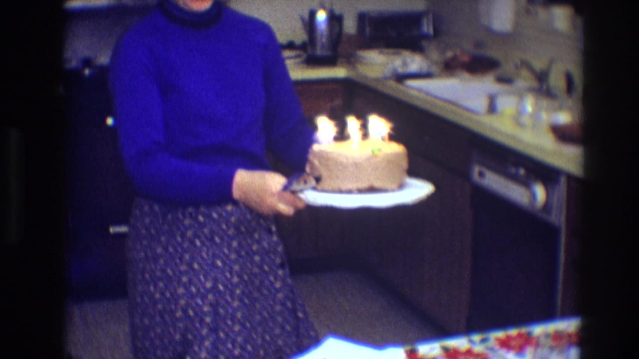 Woman carries a lit birthday cake on a white plate past the kitchen counter.